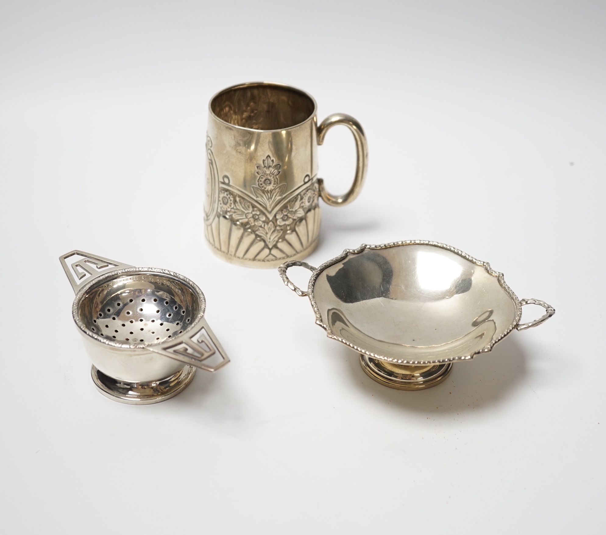 An Edwardian embossed silver christening mug, Birmingham, 1903, height 8cm, together with a silver tea strainer on stand and a small silver two handled dish, 6.9oz.